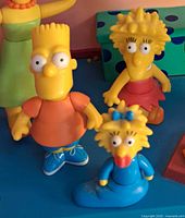 Four Simpsons family character figures including Bart, Lisa, Maggie, and Marge shown against playset background