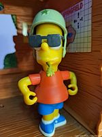 Close-up of Bart figure inside the tree house wearing a green helmet with star, sunglasses, orange shirt, blue shorts, and blue shoes with a green sprig in mouth.