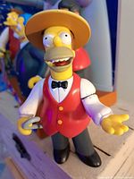 Close-up of Homer Simpson figure in red vest and yellow hat, showing detailed facial features and attire to confirm authenticity and condition.