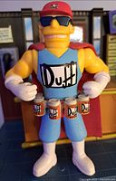 Close-up of Duff Man action figure with red cap, sunglasses, cape, and belt holding four Duff beer cans.