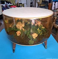 Front view of the large inflatable terrarium footstool, showing amber-tinted plastic body with artificial flower arrangement inside, and white padded top supported by three wooden legs.
