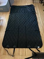 Full view of black quilted car liner laid flat on wooden floor showing straps and buckles.