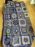 Photo of full handmade crochet blanket showing all square crochet patterns in various color combinations of purple, blue, green, cream and yellow, laid out on a floor.