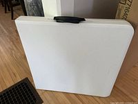 White folding table folded flat with built-in handle visible, showing portability.