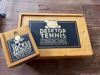 Wooden rectangular box labeled 'Desktop Tennis' and smaller square box labeled 'Jacks Osselets' on wooden floor surface.