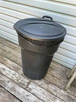 Full view of black large round garbage can with lid and wheels, placed outside on wooden deck against white siding.