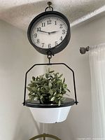 Full view of the hanging clock with visible black metal frame, white enamel bowl, and artificial plant inside bowl.