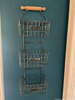 Full view of hanging kitchen shelf with three wire baskets and wooden rolling pin style top handle against a dark blue wall.