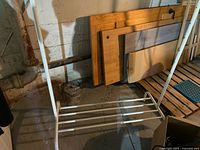 White metal rolling clothing rack frame showing lower shelf with parallel metal rods against a background of wooden planks and a concrete wall.
