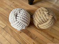 Two spherical rope door stoppers placed on a wood floor, one tan and one white, showing their shape and texture.