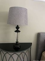 Table lamp with glass body and dark mirrored base, paired with grey cylindrical fabric lampshade on a black half-moon decorative table.