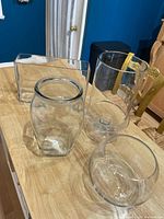 Four clear glass vases shown on a wooden table, including rectangular, cylindrical, twist, and round shapes.