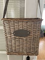 Front view of wicker basket with visible woven pattern, metal 'WELCOME' plaque and dark hanging strap attached.