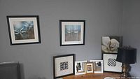 Framed artwork on grey wall with table below containing smaller framed artworks and a lamp