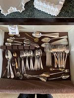 Top view of assorted silver plated flatware laid out in a tray. Includes forks, spoons, and serving utensils with decorative handles and some patina visible.