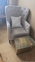 View of the upholstered wingback chair with light gray fabric, button tufting, and floral cushion, next to the brass wood box