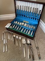 Complete silver plate flatware set displayed in open blue fabric-lined wooden case with several pieces laid outside the case on carpeted floor.