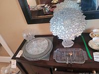 View of multiple crystal and art glass dishes arranged on a dark wooden table beneath a mirror, including a large iridescent art glass plate on a stand, crystal round plates and bowls, and a rectangular serving tray with silver utensils.