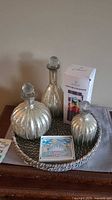 Overall lot showing three silver-tone ribbed glass decanters on woven basket tray with decorative tile and boxed portable blender.