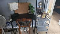Desk with lamp, cork board, clock, chair with floral upholstery, metal rolling shelf with book on top