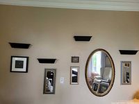 Wide view of the wall showing all items mounted including oval mirror, 2 framed rectangular mirrors, small framed art, and 3 black wall shelves.