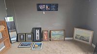 Wide shot showing all framed wall art and door mirror arranged on floor against wall, including various framed prints and original painting, plus door mirror.