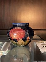 Front view of the vase showing the large red fruit and surrounding smaller fruits with leaves against the dark background.