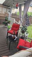 Wide view of patio set showing two black metal chairs with red cushions, small side table, plants, and decorative bird centerpiece on a covered wooden deck with curtains.