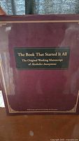 Close up of the large burgundy hardcover book titled 'The Book That Started It All: The Original Working Manuscript of Alcoholics Anonymous'