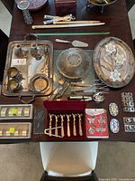 Top-down image showing two silver plated trays, assorted tableware items including spoons in a red case, salt and pepper shakers, a metal serving bowl, and various serving utensils.
