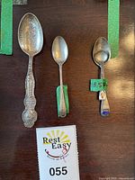 Three silver spoons laid on wooden surface: from left to right large engraved Olympic Range Seattle teaspoon, plain Demitasse spoon, salt spoon with two enamel crests on handle.