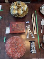 Overview of all items: gold painted fruit-shaped gas candles in brass bowl, pair of beige chopsticks, chopstick rest, decorative wooden box, carved wooden bowl with flower design, small candle holder, silver serving utensils.
