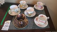 All six teacups each on matching saucers displayed with one extra saucer, arranged on a green table surface.