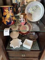 Shelf display with Tunstall ceramic vase and jug, floral decorative plates, hobnail milk glass vase, ashtrays, bowl, and crystal bowl and glass with red accents.