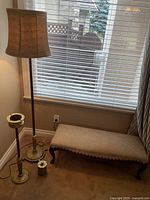 Photo showing vintage brass floor lamp, standing ashtray, brass cigarette box, and wood carved cushioned bench in room corner