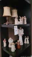 Photo of multiple wood carved angel figurines placed on two shelves of a black shelving unit, along with two ornate table lamps.