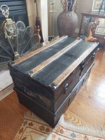Antique black wooden steamer trunk with metal bands, corner protectors, and central locking latch displayed on hardwood floor near fireplace.