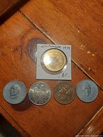 Six Canadian collectible coins and medals arranged on a table, showing variety of designs including trade dollar in holder and tokens with provincial and floral motifs.