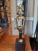 Full view of brass eagle pedestal wick and fluid lantern lighter on wooden surface, showing detailed metalwork and stone base.
