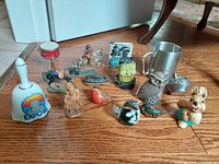 Group image of various collectibles including Expo 86 bell, pewter cup, birds, Spongebob, bunny, drum, and more on wood floor.