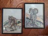 Two framed wildlife art pieces by E Warner depicting a bear and a ram, placed side by side on a wooden floor.