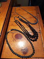 Three vintage stone necklaces displayed on wooden surface: one single strand of smooth hematite beads, one triple strand of black faceted beads, and one strand of marbled green beads.