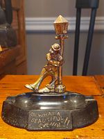 Front view of black bakelite ashtray, showing man figurine leaning on street lamp and engraved text on front