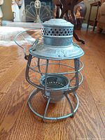 Metal lantern cage with wireframe protective cage and ventilated top viewed from the side on wood floor.