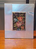 Front and angled views of the rectangular vintage compact showing mother of pearl inlay and central petit point embroidery floral panel framed in gold plating.