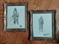 Two bronze metal ornate framed black and white sketches of figures, under glass, on wood floor.