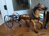 Front-side angle showing horse and sulky cart with metal frame and wooden seat on wooden floor.