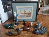 Photo showing six hand carved and painted wood bird sculptures arranged on wood floor alongside a framed waterfowl print in wood frame with glass