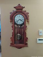 Front view of vintage mechanical key wind pendulum chiming wall clock showing wood casing, clock face with Roman numerals, and pendulum behind glass door with ornamental golden painted designs.