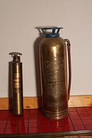 Two antique brass fire extinguishers standing side by side on a tiled floor against a wall.
